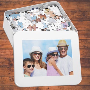 Peut inclure: Une bo&icirc;te de puzzle blanche avec une photo de famille sur le devant. La photo montre une famille de quatre personnes souriantes et portant des chapeaux. La bo&icirc;te de puzzle est ouverte et les pi&egrave;ces du puzzle sont visibles &agrave; l'int&eacute;rieur.