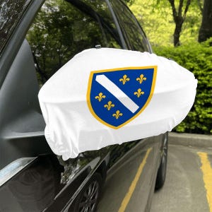 May include: White car mirror cover displaying the coat of arms of Bosnia and Herzegovina. The design features a blue shield with a white diagonal stripe and six gold fleur-de-lis. The cover is fitted to a black car.