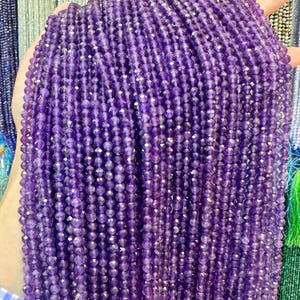 Könnte beinhalten: Stränge aus facettierten, tiefvioletten Amethystperlen. Die kleinen, runden Perlen haben eine polierte, glitzernde Oberfläche. Die Stränge werden zusammengehalten, wodurch eine dichte, strukturierte Darstellung des Edelsteins entsteht.