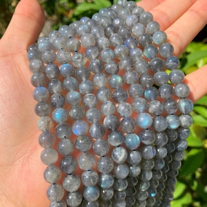 Könnte beinhalten: Strang aus grauen Labradorit-Edelsteinperlen. Die runden Perlen haben einen Durchmesser von etwa 8 mm. Sie weisen eine subtile Irisierung mit blauen und grünen Reflexen auf. Die Perlen sind auf einer blauen Schnur aufgefädelt.