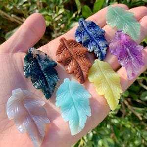 Könnte beinhalten: Eine Sammlung von geschnitzten Edelstein-Blattanhängern in verschiedenen Farben, darunter Grün, Blau, Lila, Gelb und Braun. Jedes Blatt ist etwa 5 cm lang und zeigt detaillierte Adern und Texturen. Die Anhänger werden in einer Hand gehalten.