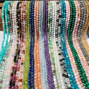 May include: A variety of gemstone beads in different colors and textures, including turquoise, amethyst, green aventurine, and rose quartz. The beads are strung on cords and displayed in rows.