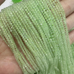 以下が含まれることがあります： 薄緑色のファセットビーズのストランドのクローズアップ。ビーズは小さく、一緒にひもでつながれており、きらめく効果を生み出しています。ビーズは手で持たれており、ストランドが滝のように流れ落ちています。全体的な色は、柔らかいパステルグリーンです。