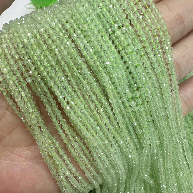 以下が含まれることがあります： 薄緑色のファセットビーズのストランドのクローズアップ。ビーズは小さく、一緒にひもでつながれており、きらめく効果を生み出しています。ビーズは手で持たれており、ストランドが滝のように流れ落ちています。全体的な色は、柔らかいパステルグリーンです。
