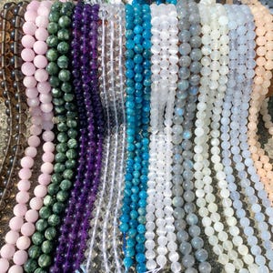 Può includere: Una collezione di fili di perline in vari colori, tra cui rosa, verde, viola, trasparente, blu, bianco, grigio e pesca. Le perline rotonde sembrano essere di pietra o cristallo lucido, adatte per la creazione di gioielli o l'artigianato.