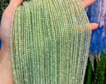 May include: A handful of light green gemstone beads. The beads are small, round, and strung together in long strands. The beads have a faceted cut, giving them a sparkling appearance. The background is blurred, with hints of other colors.