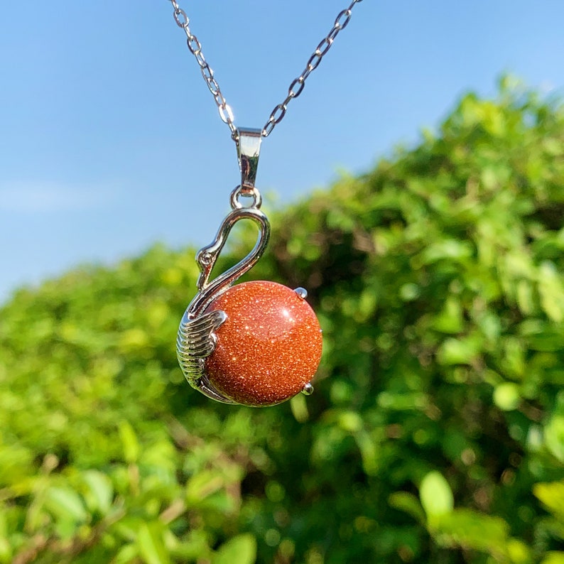 Pode incluir: Um colar de prata com um pingente em forma de cisne segurando uma pedra de sol redonda e laranja.