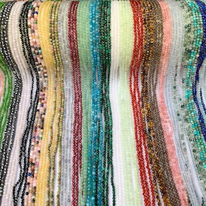 Könnte beinhalten: Eine Auswahl an Edelsteinperlen in verschiedenen Farben und Formen, darunter Schwarz, Weiß, Grün, Blau, Rot und Braun. Die Perlen sind auf einem weißen Hintergrund aufgereiht.