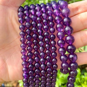 Könnte beinhalten: Nahaufnahme von mehreren Strängen tiefvioletter Amethystperlen. Die kugelförmigen Perlen sind aufgereiht und zeigen eine satte, durchscheinende Farbe. Die Perlen werden in einer Hand gehalten, was darauf hindeutet, dass sie für die Schmuckherstellung oder Präsentation bereit sind.