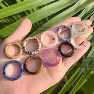 Op de afbeelding: Een verzameling edelsteenringen in verschillende kleuren, waaronder groen, roze, blauw en zwart. De ringen worden in een hand gehouden, waarbij hun verschillende texturen en patronen worden getoond. De ringen zijn gemaakt van verschillende soorten stenen.