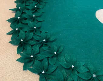 May include: A green felt Christmas tree skirt with a glittery finish. The skirt is decorated with a row of green felt poinsettia flowers with white pearl centers.