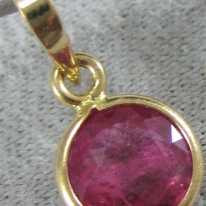 May include: A gold pendant with a round, bezel-set ruby gemstone. The ruby is a deep pink color and has a faceted cut.