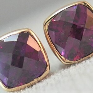 May include: A pair of square-cut, faceted, deep purple gemstone stud earrings with gold-colored settings. The earrings have a cushion-like shape and reflect light, showcasing their multiple facets. The gold settings provide a warm contrast to the dark purple gems.