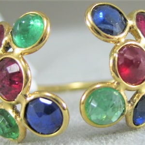 Puede incluir: Un anillo de oro con un grupo de piedras preciosas redondas en rojo, verde y azul. El anillo está abierto por un lado y tiene una forma curva.