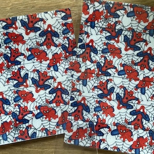 Könnte beinhalten: Zwei rechteckige Bögen aus Kunstleder mit einem sich wiederholenden Muster des Superhelden Spider-Man in rot-blauen Anzügen, dargestellt in verschiedenen Posen vor einem Hintergrund aus schwarzen Spinnennetzen auf weißer Fläche.