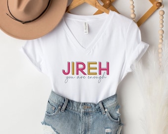 Può includere: Maglietta bianca a collo a V con il testo "JIREH you are enough" in lettere colorate. La maglietta è appesa a un appendiabiti di legno con un cappello di paglia e un vaso di erba della pampa secca.