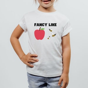 May include: White t-shirt with a red apple and bees graphic and the text "FANCY LIKE" printed on the front.