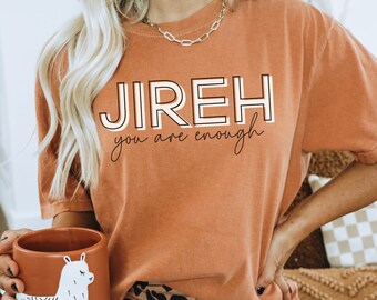 Può includere: Una donna indossa una maglietta color arancio bruciato con il testo "JIREH you are enough" in bianco e nero. Indossa anche pantaloncini leopardati marroni e neri e tiene in mano una tazza marrone con un disegno di lama bianco.