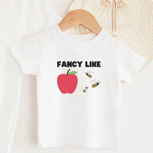 Puede incluir: Camiseta blanca de manga corta con una manzana roja y el texto "FANCY LIKE" en negro. Hay cuatro abejas amarillas y negras volando alrededor de la manzana.