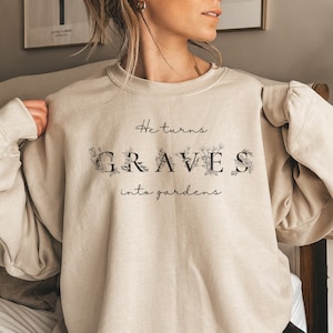 Może przedstawiać: Beżowa bluza z nadrukiem "He turns GRAVES into gardens" na przodzie. Tekst jest otoczony wzorami kwiatowymi.
