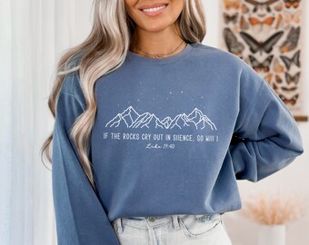 Puede incluir: Una sudadera azul con un gráfico de una cordillera de montañas blancas y el texto "IF THE ROCKS CRY OUT IN SILENCE, SO WILL I" y "Luke 19:40".