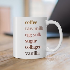 May include: White ceramic mug with the words "coffee, raw milk, egg yolk, sugar, collagen, vanilla" printed in brown and pink.