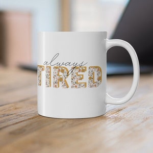 May include: White ceramic coffee mug with a floral design. The text "always tired" is printed on the mug in black and gold.