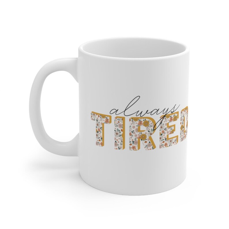 May include: White ceramic mug with a floral pattern in gold and pink. The text "always tired" is printed on the mug in a gold font.
