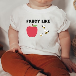 May include: A white baby t-shirt with the text "FANCY LIKE" printed in black. A red apple and four yellow and black bees are printed on the shirt.