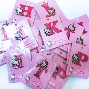 Peut inclure: Une collection de patchs thermocollants roses représentant les lettres de l'alphabet et un personnage Hello Kitty. Les patchs sont sur des cartes roses avec le texte "Alphabet Iron-on Patch".