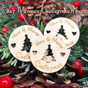 Può includere: Tre ornamenti natalizi in legno con un'incisione di un albero di Natale e il testo "Have a Merry Christmas" su ciascuno. Gli ornamenti sono su un letto di rami di pino verdi e bacche rosse.