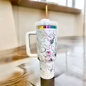 May include: White travel mug with a handle and a straw. The mug features a colourful, iridescent design with Mickey Mouse, a castle, and other Disney-themed icons. The text "Magic" is visible on the mug.