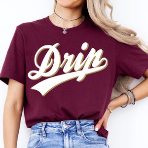 May include: Burgundy crew neck t-shirt with the word "Drip" in a white script font with a gold outline. The shirt is made of a soft material and is suitable for casual wear.