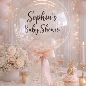 May include: A clear balloon with the text "Sophia's Baby Shower" in black script, filled with pink confetti and smaller balloons. The balloon is tied with a pink bow and ribbon, part of a baby shower theme with pink and white decorations.