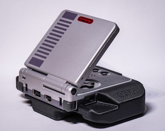 May include: A silver Game Boy Advance SP with a black and gray case. The Game Boy Advance SP is open and the screen is visible. The screen has a gray background with a black Nintendo logo. The case has the word "GETA" printed on it.