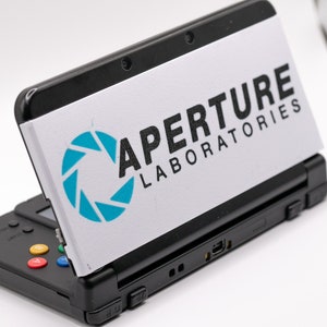 May include: A black Nintendo 3DS handheld gaming console with a white custom faceplate featuring the Aperture Laboratories logo and a blue and white aperture science logo.