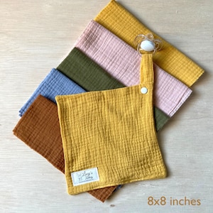 Peut inclure: Un ensemble de six langes en mousseline pour bébé de 20,3 x 20,3 cm dans différentes couleurs, dont le jaune moutarde, le marron, le vert, le bleu et le rose. Les langes ont une fermeture à pression et une boucle pour attacher une sucette. Les langes sont fabriqués par Lucy's Little Shop.