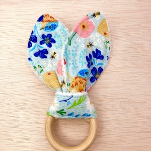 May include: A light blue fabric baby teether with a floral and bird print. The teether is tied around a wooden ring.