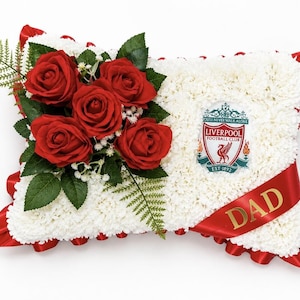 May include: A rectangular floral pillow arrangement made of white carnations with a red ribbon border. It features a cluster of four red roses with green leaves and a fern, the Liverpool Football Club logo, and a red sash with the word DAD in gold.
