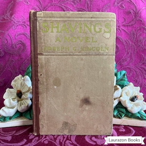 May include: A vintage book titled "Shavings, A Novel" by Joseph C. Lincoln. The book has a tan cover with green lettering and a brown spine. The book is propped up by a white floral bookend on a patterned purple background.