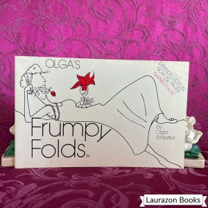 May include: A rectangular book titled Olga's Frumpy Folds by Olga Schpiffeir. The white cover features a black line drawing of a person reclining, holding a red flower in their mouth and a red napkin fold in a glass. Text reads Expanded Edition Now 112 Pages.