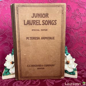 May include: A vintage book titled "Junior Laurel Songs" with a special edition label. The book is brown with a dark brown border and the title is printed in black. The author is listed as M. Teresa Armitage and the publisher is C.C. Birchard & Company, Boston.