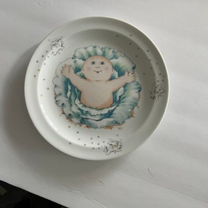 May include: White ceramic plate featuring a baby design. The baby is surrounded by a blue and green cabbage-like design. The plate also has illustrations of small bunnies and stars.