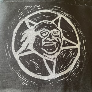 May include: A black and white patch with a white pentagram and a white image of a person wearing glasses inside the pentagram. The patch is sewn onto a black fabric.