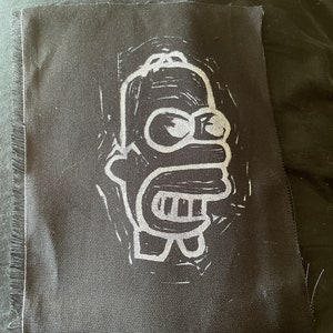 May include: A black and white embroidered patch of a cartoon character with a yellow face and blue hair. The character is wearing a white shirt and has a large smile.