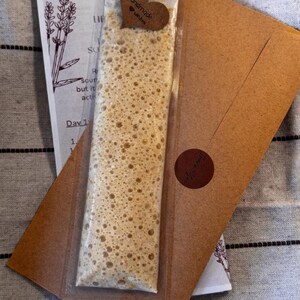 May include: A package containing a light brown, bubbly substance in a clear plastic sleeve, with a heart-shaped tag that reads "Handmade with love." The package rests on a brown envelope and a card with lavender illustrations.