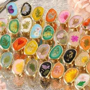 Op de afbeelding: Een verzameling ovale ringen met goudkleurige banden, elk met een andere gedroogde bloem of plantenmonster ingekapseld in heldere hars. De ringen zijn gerangschikt op een zilveren decoratief dienblad, met roze bloemen op de achtergrond.