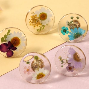 May include: Five clear resin rings, each with a different dried flower arrangement. The flowers include white daisies, yellow daisies, purple flowers, and blue flowers. The rings are on a yellow and pink background.