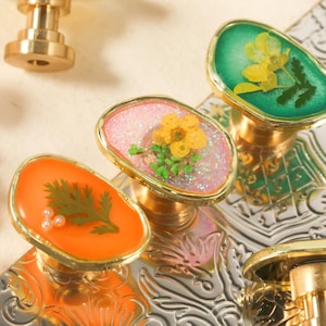 May include: Close-up of decorative cabinet knobs with gold-colored bases and oval tops. The tops are filled with colorful resin and dried flowers. The knobs are in shades of orange, pink, and green.