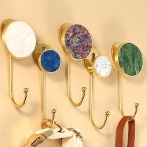 Puede incluir: Un conjunto de cinco perchas de pared decorativas con herrajes metálicos dorados. Cada gancho presenta un elemento decorativo circular en blanco, azul, estampado, blanco y verde. Los ganchos están montados en una pared beige claro, con un collar para perro y una correa de cuero marrón.